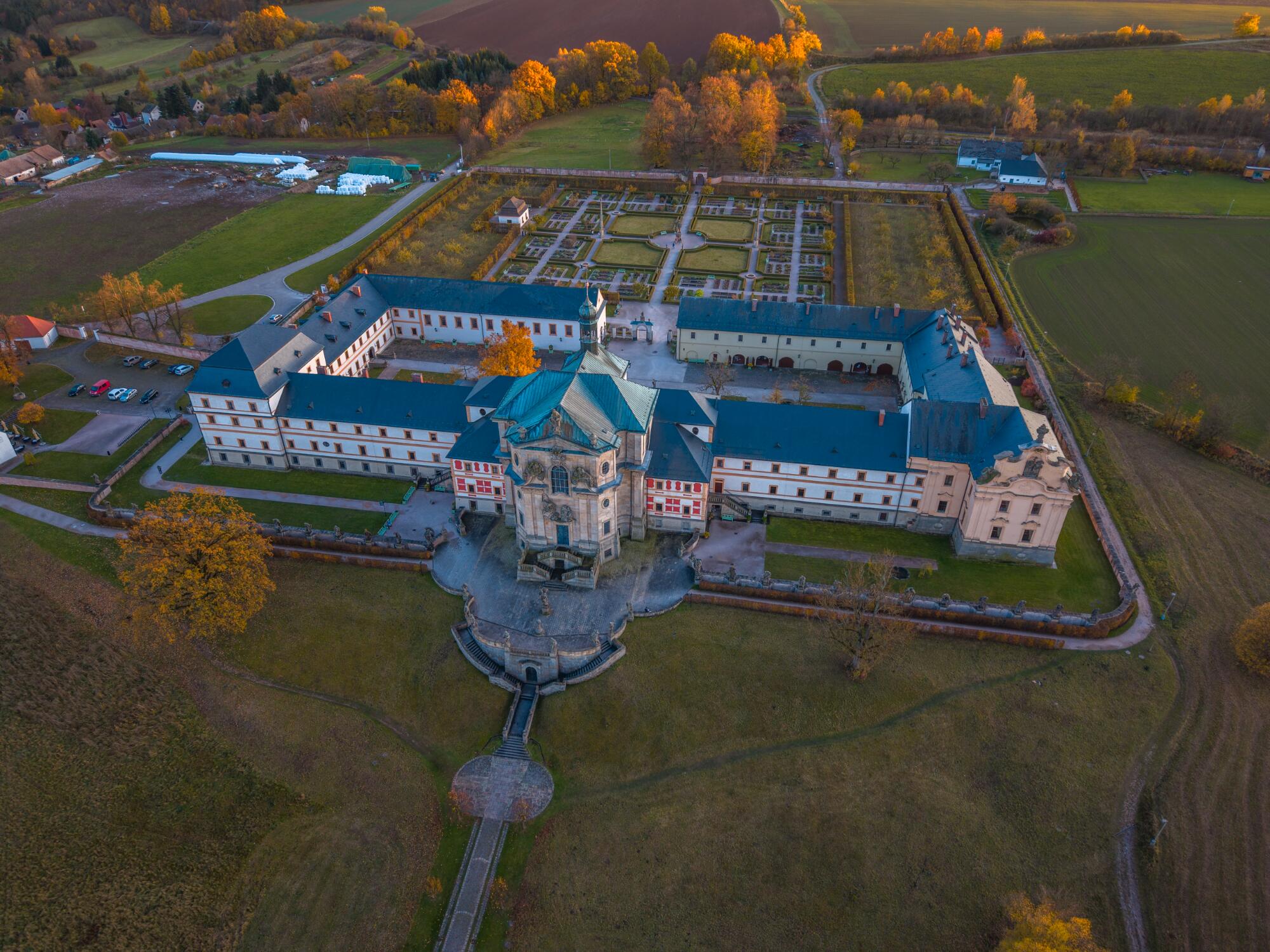 Preview of: Kuks Spa and Hospital Vue aérienne d’un vaste ensemble architectural avec cour et jardins géométriques, entouré de champs, d’arbres et de chemins ruraux.
