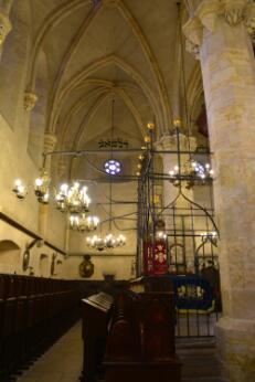 Intérieur d'une synagogue médiévale voûtée, avec arche sainte grillagée, bancs de prière et luminaires suspendus.