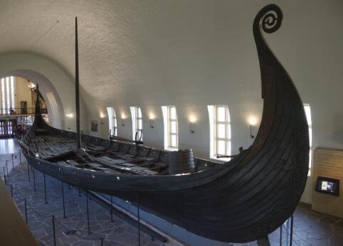 Navire viking en bois exposé dans un musée, caractérisé par sa coque élancée et sa proue sculptée recourbée.