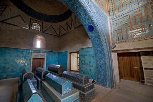 Photographie représentant l'intérieur d'un mausolée, avec plusieurs sarcophages recouverts de carreaux turquoise sous une architecture voûtée ornée de décors géométriques.