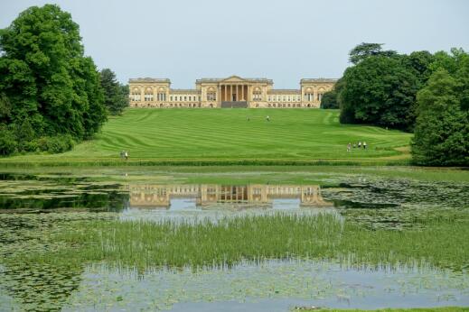 Vue d'un vaste jardin paysager structuré autour d'une grande pelouse et d'un bassin, dominé par un palais de style néoclassique.