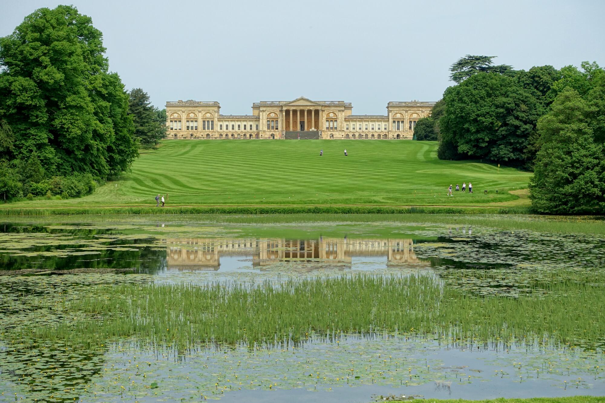 Preview of: Stowe Gardens Vue d'un vaste jardin paysager structuré autour d'une grande pelouse et d'un bassin, dominé par un palais de style néoclassique.