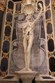 Photographie représentant une statue funéraire en marbre figurant un squelette debout levant un bras, placée dans une niche richement décorée.