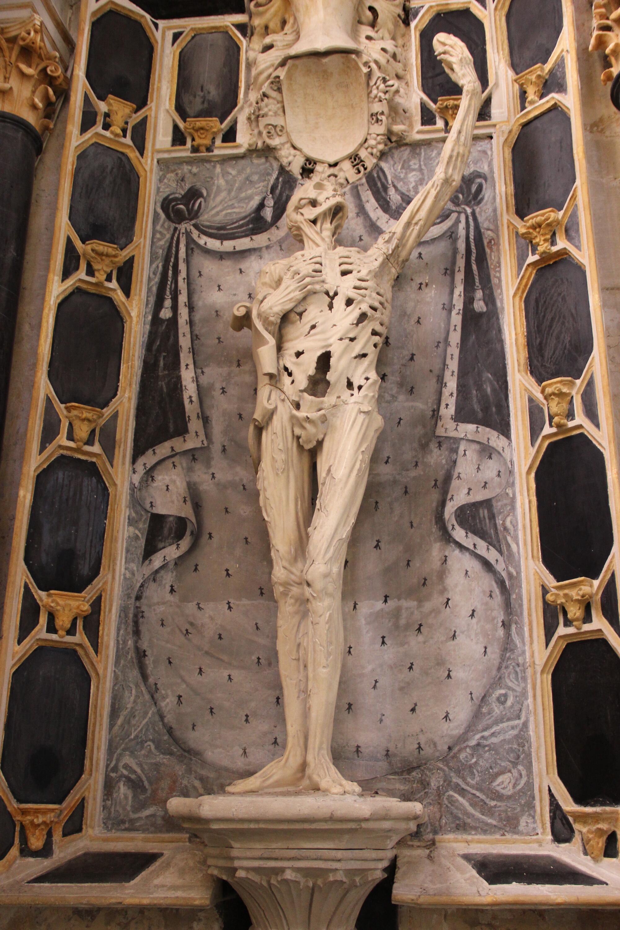 Preview of: Transi (Cadaver Tomb) of René of Chalon Photographie représentant une statue funéraire en marbre figurant un squelette debout levant un bras, placée dans une niche richement décorée.