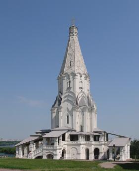Église blanche à haute flèche centrale, entourée de galeries et d’escaliers, située sur une pelouse sous un ciel bleu dégagé.