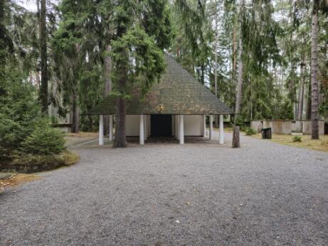 Petit bâtiment à toit pyramidal soutenu par des colonnes, situé dans une clairière entourée de grands arbres, avec une allée de gravier au premier plan.