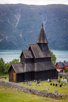 Église en bois debout de style médiéval, à toiture à plusieurs niveaux et clocher central, entourée d'un cimetière et située au bord d'un fjord montagneux.