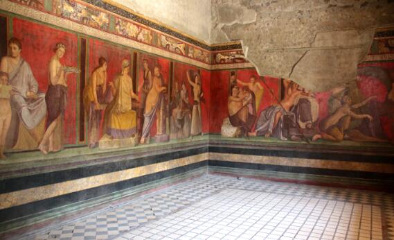 Salle antique ornée de fresques murales polychromes représentant des personnages dans des scènes rituelles ou mythologiques, avec un sol en mosaïque géométrique.