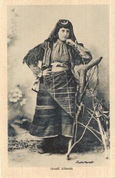 Portrait photographique d'une femme portant un costume traditionnel albanais composé d'une longue jupe, d'un tablier à motifs, d'un voile et d'un gilet brodé, posant debout dans un décor de studio.