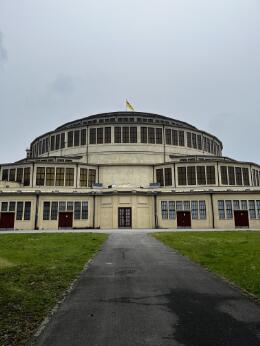 Grand bâtiment circulaire à plusieurs niveaux de fenêtres, vu de face au bout d’une allée, avec pelouses et ciel couvert.
