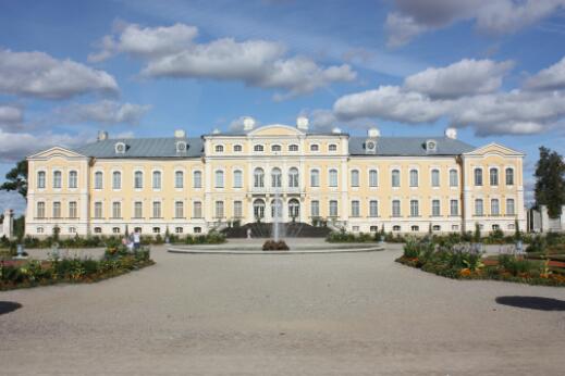 Photographie montrant la façade symétrique d’un vaste palais baroque de couleur jaune pâle, précédé d’un jardin à la française avec une fontaine centrale sous un ciel nuageux.