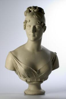 Buste en marbre représentant une jeune femme à l’expression calme et idéalisée. Elle porte une coiffure élaborée relevée en chignon, ornée de boucles encadrant le visage, et une robe drapée à large décolleté. La sculpture, de style néoclassique, est présentée sur un petit piédouche et se détache sur un fond neutre clair.