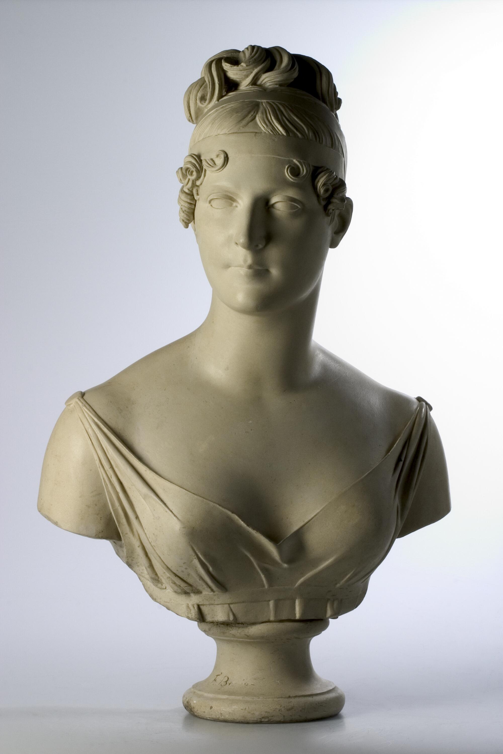 Preview of: Buste de l'impératrice Joséphine Buste en marbre représentant une jeune femme à l’expression calme et idéalisée. Elle porte une coiffure élaborée relevée en chignon, ornée de boucles encadrant le visage, et une robe drapée à large décolleté. La sculpture, de style néoclassique, est présentée sur un petit piédouche et se détache sur un fond neutre clair.