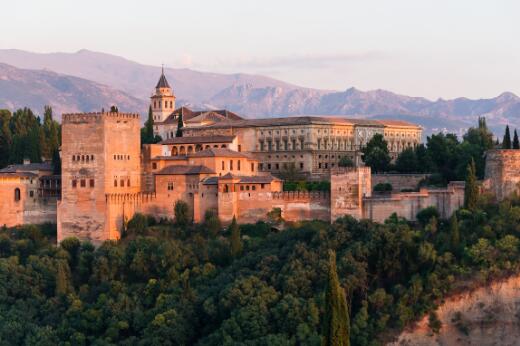 Vue panoramique de l'Alhambra de Grenade, vaste ensemble palatial et fortifié dominant une colline boisée, avec ses tours, remparts et palais se détachant sur fond de chaîne montagneuse au coucher du soleil.