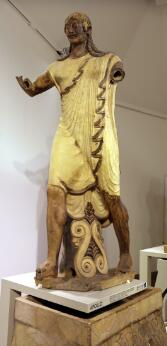Statue en terre cuite polychrome représentant une figure masculine debout vêtue d'une tunique drapée, les bras partiellement conservés, reposant sur un socle orné de motifs en volutes.